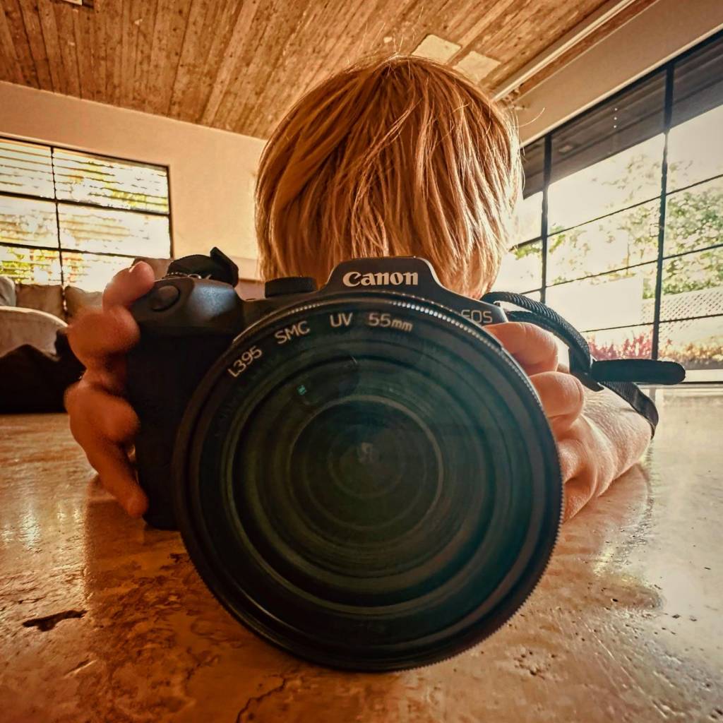 ילד אוחז מצלמת קנון גדולה ומביט דרכה, צילום ביתי באור טבעי על רצפת שיש עם רקע חמים